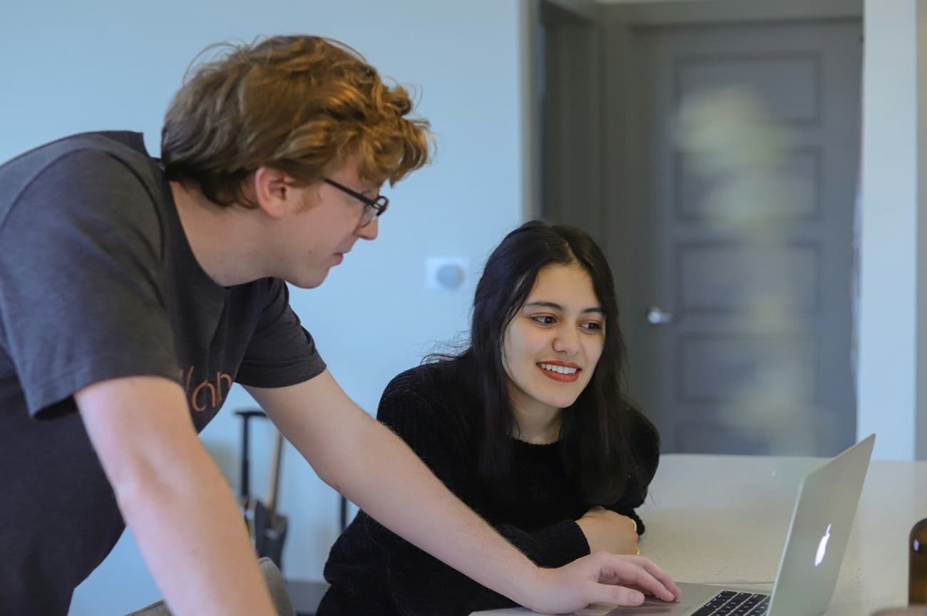 Someone leaning in to explain something on screen to a student at a laptop